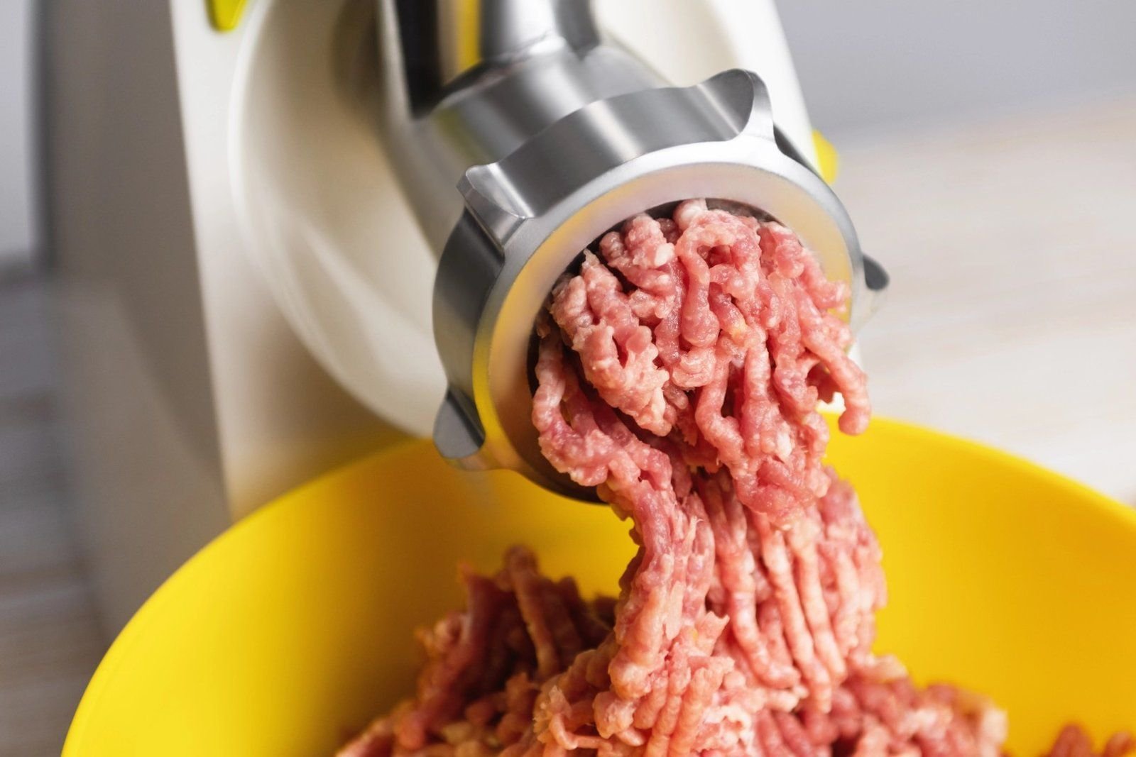 Freshly ground raw beef coming out of a stainless steel meat grinder into a yellow bowl.
